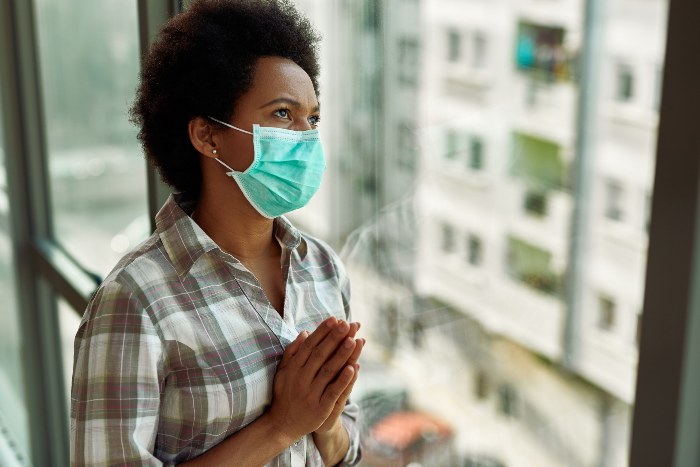 African American woman with protective face mask praying by the window.